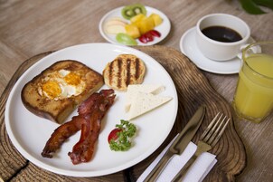 Un plato de desayuno con tostadas, tocino, queso y una ensalada pequeña, una taza de café y un vaso de jugo de naranja sobre una mesa de madera.