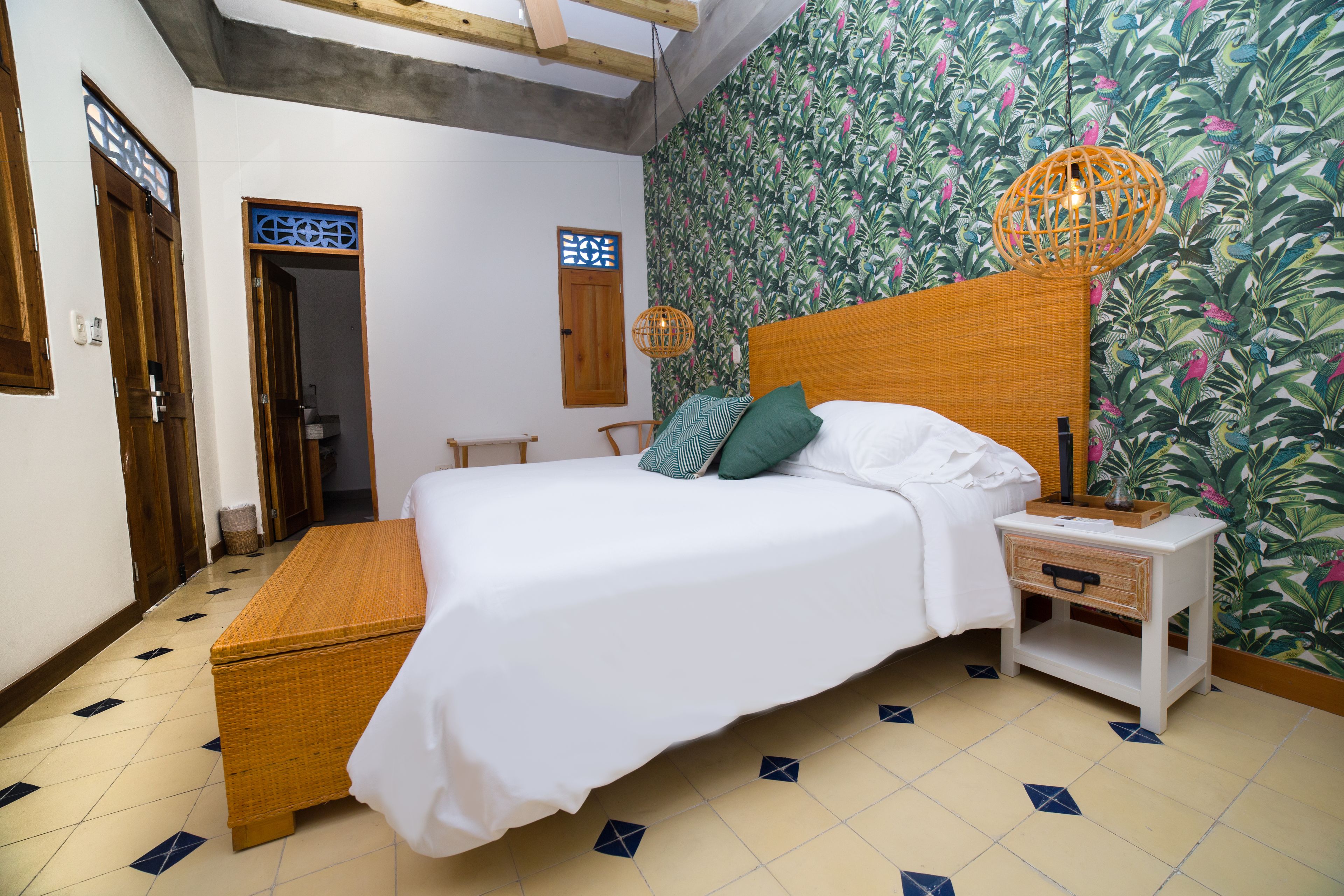 A bedroom with a bed, a nightstand, a wicker chest, and a wall with a tropical wallpaper.