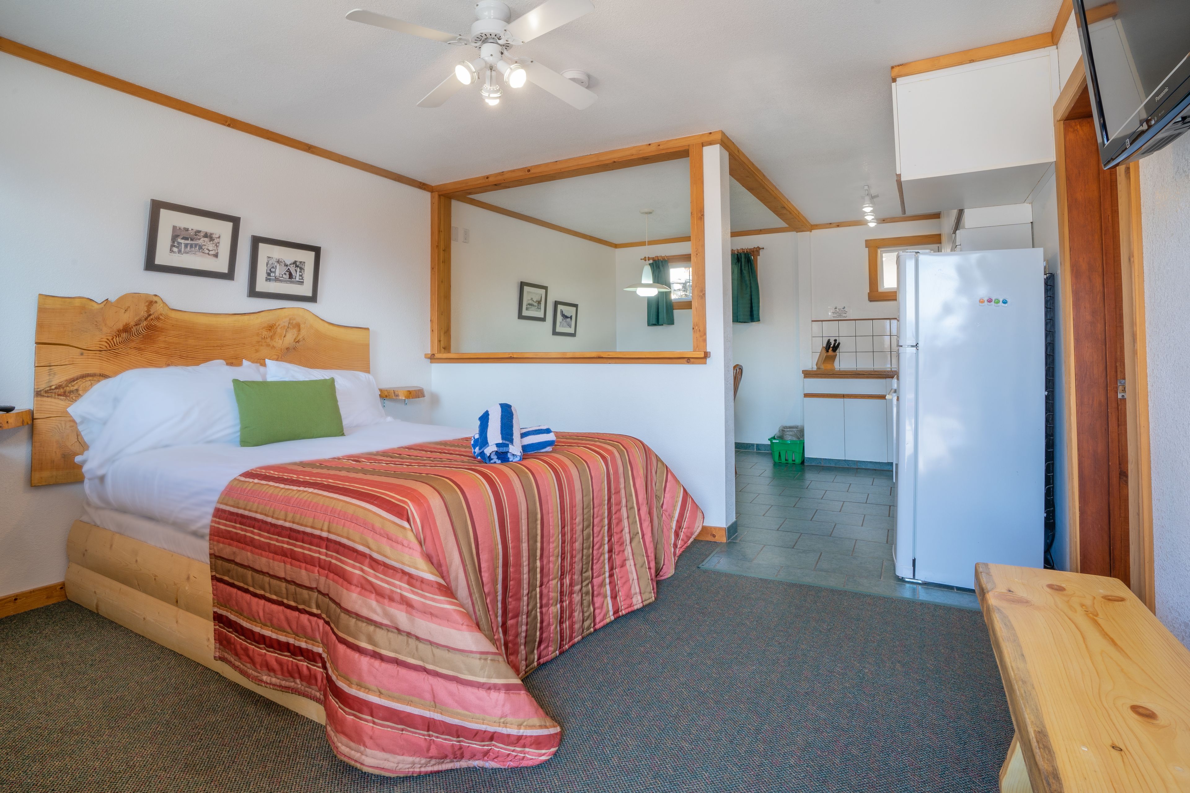 A bedroom with a bed, a wooden headboard, a mirror, a ceiling fan, and a kitchenette with a refrigerator.