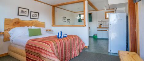 A bedroom with a bed, a wooden headboard, a mirror, a ceiling fan, and a kitchenette with a refrigerator.