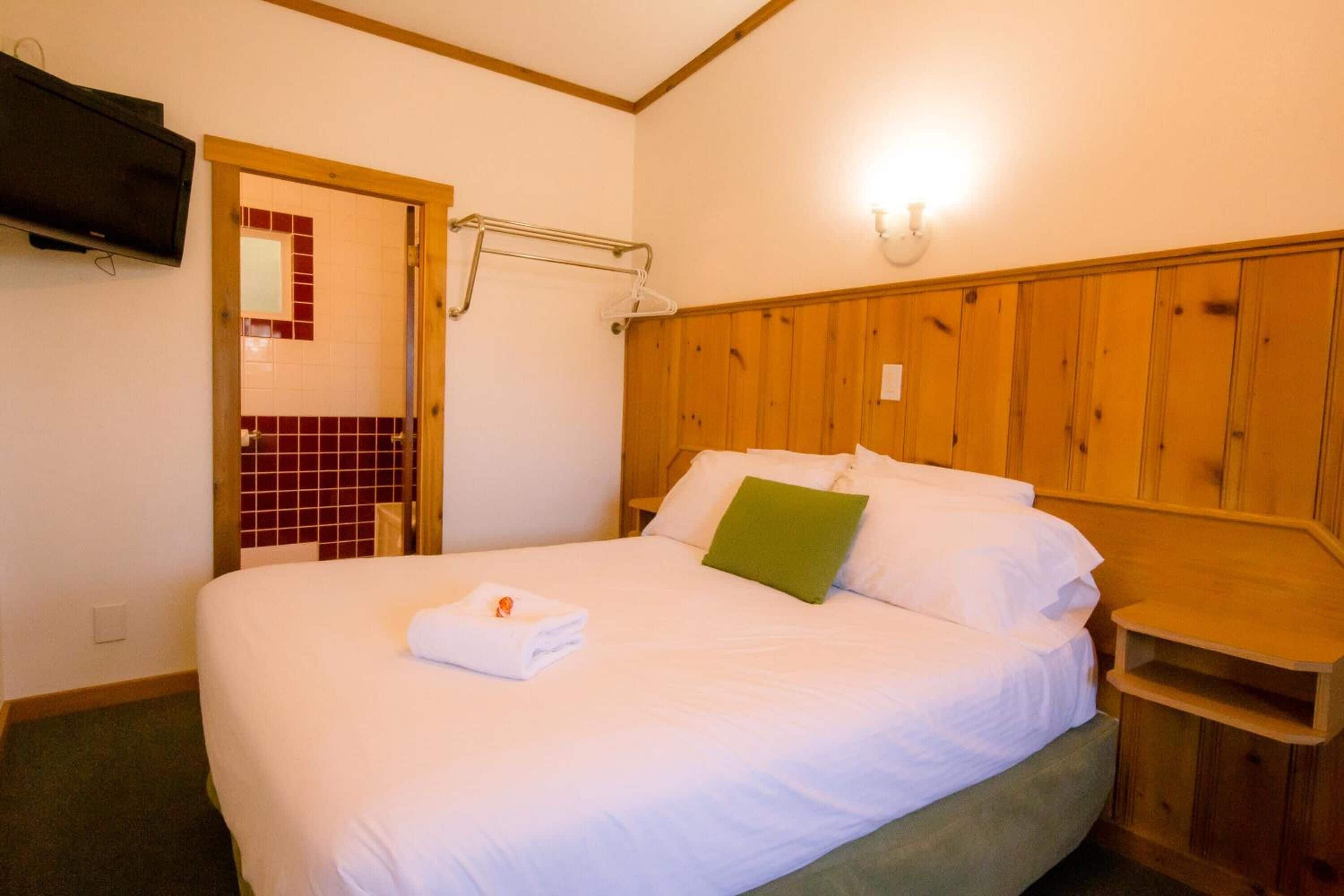 Een keurig opgemaakt bed met witte lakens en een groen kussen, een houten hoofdbord, een nachtkastje van hout, een aan de muur bevestigde televisie en een douchegedeelte met rode tegels.