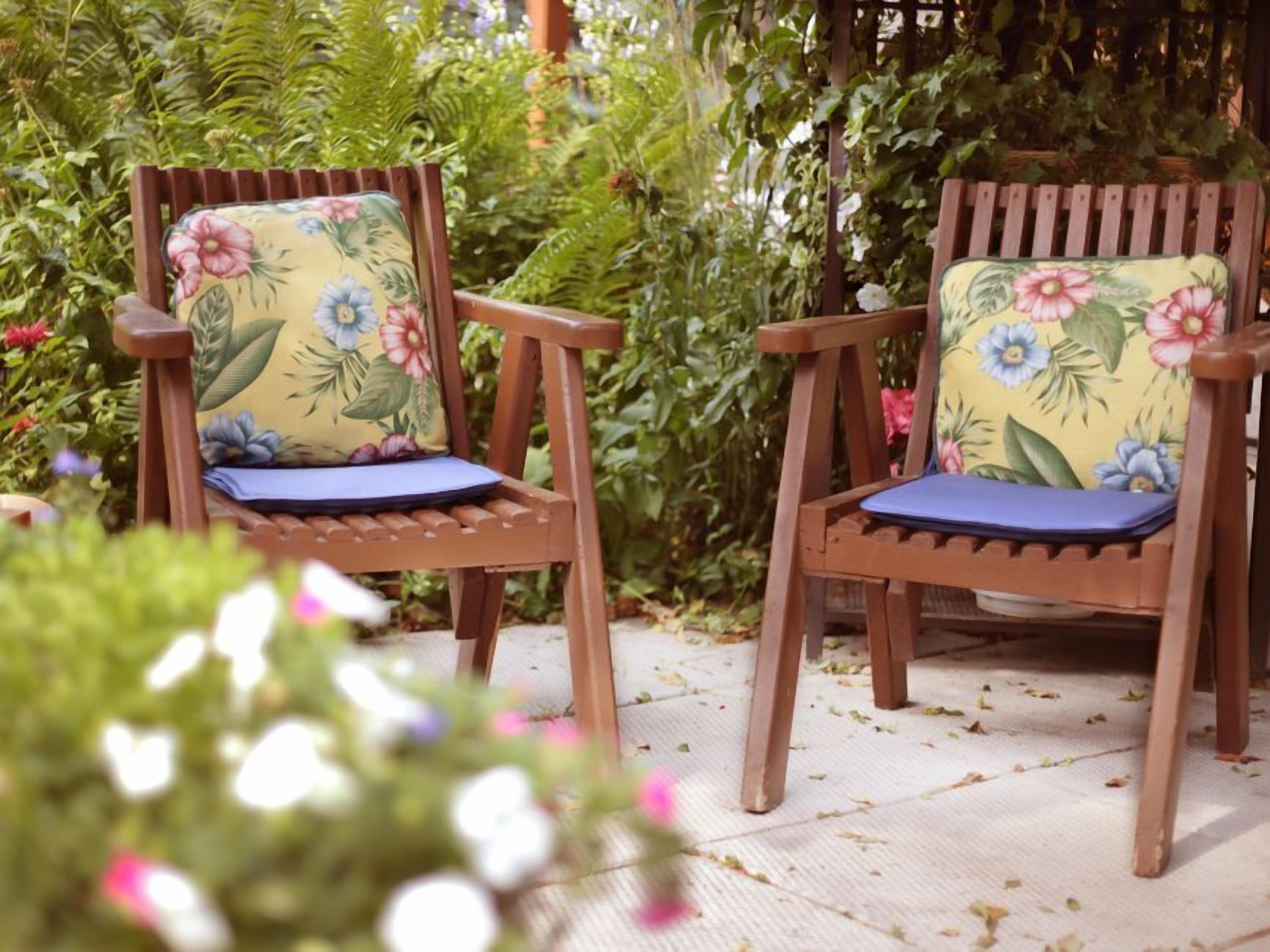 Two wooden chairs with floral cushions and a white mat on a wooden deck surrounded by greenery.