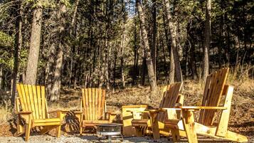 A row of wooden Adirondack chairs arranged around a metal fire pit in a forested area.