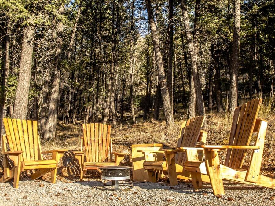 A row of wooden Adirondack chairs arranged around a metal fire pit in a forested area.