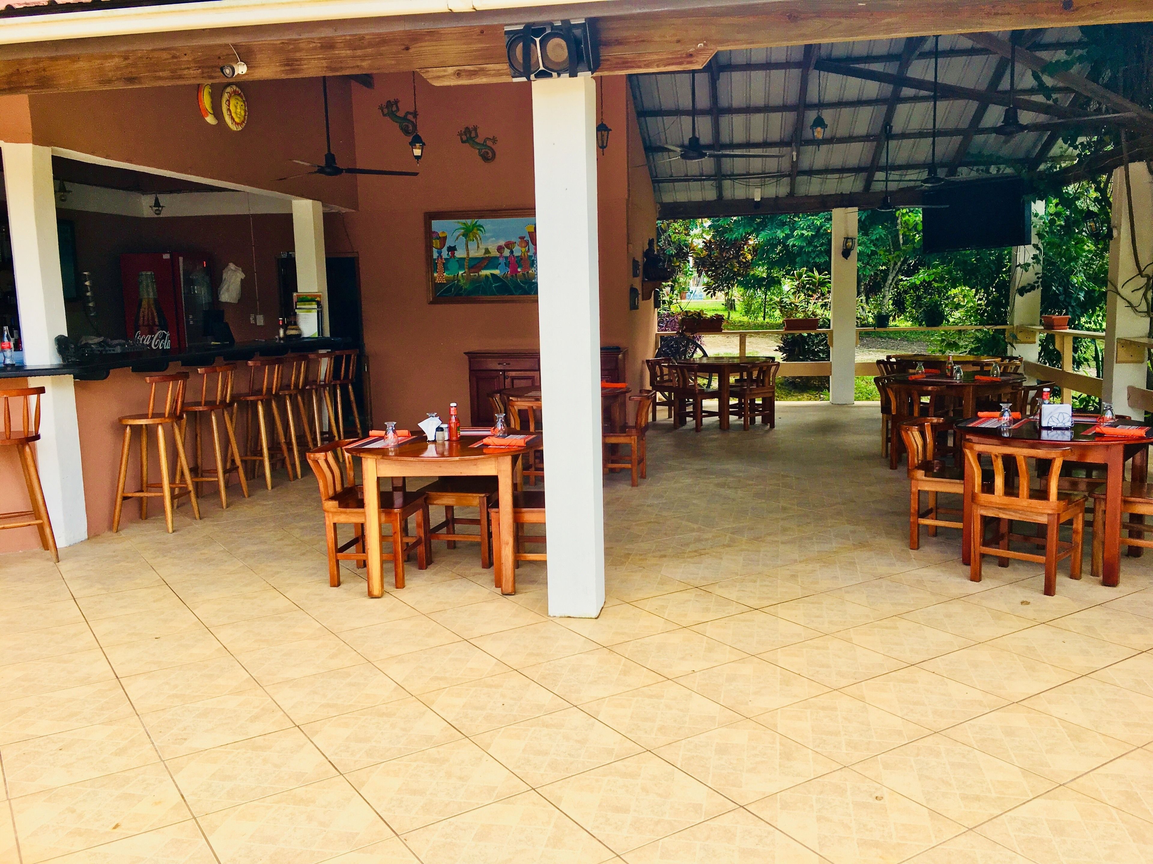 Un espacio de comedor al aire libre techado con mesas y sillas de madera, una zona de bar con taburetes y vistas a la vegetación.
