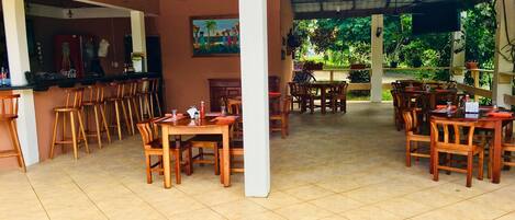 Un espacio de comedor al aire libre techado con mesas y sillas de madera, una zona de bar con taburetes y vistas a la vegetación.