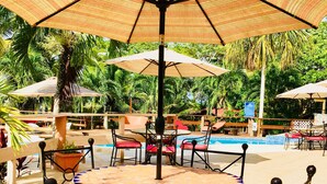 A round outdoor table with red cushioned chairs, surrounded by large umbrellas, set against a backdrop of palm trees and a pool.