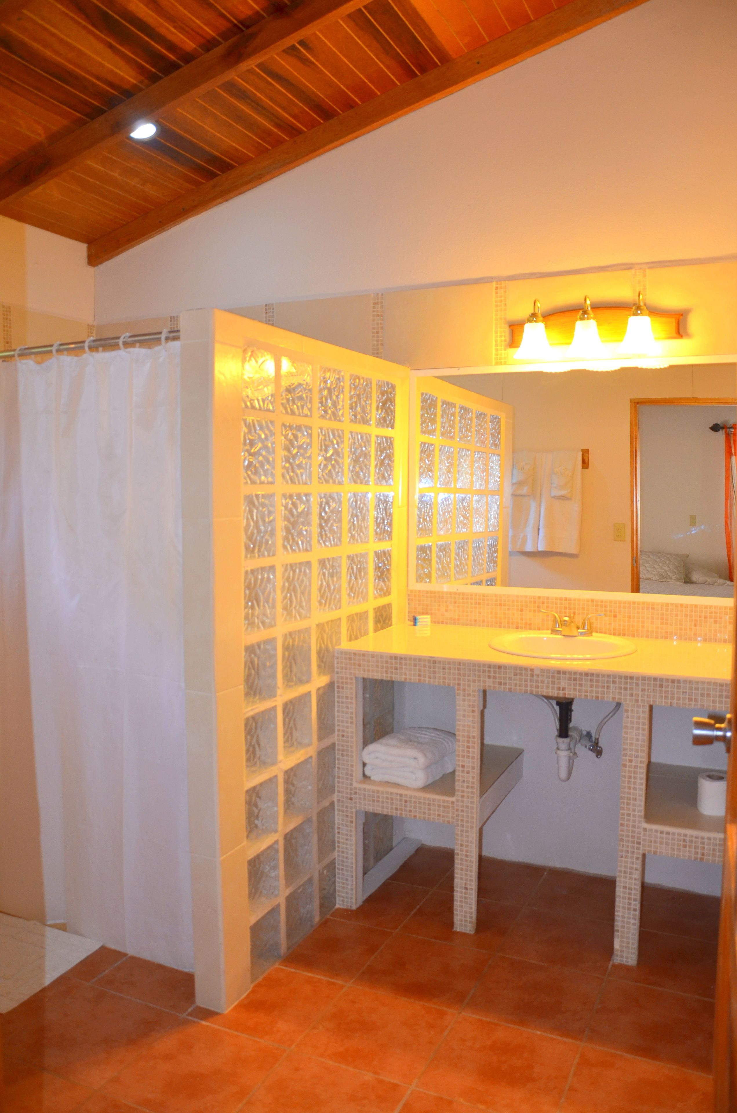 Un baño con techo de madera, cortina de ducha, lavabo con espejo y piso de baldosas.