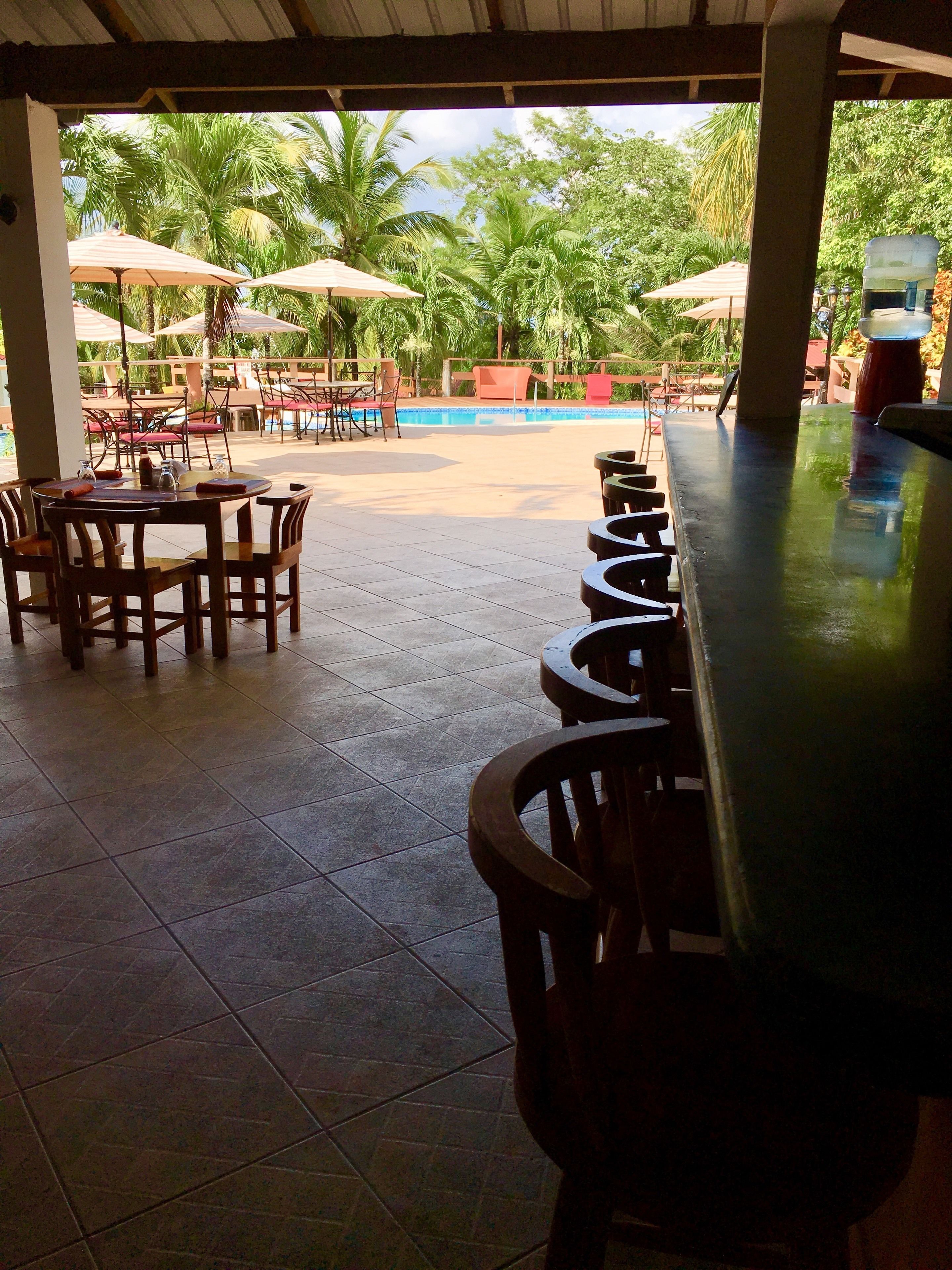 An outdoor seating area with tables and chairs, a pool, and palm trees in the background.