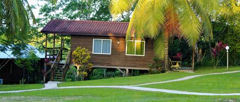 Una casa de madera con techo rojo, rodeada de abundante follaje y una palmera.