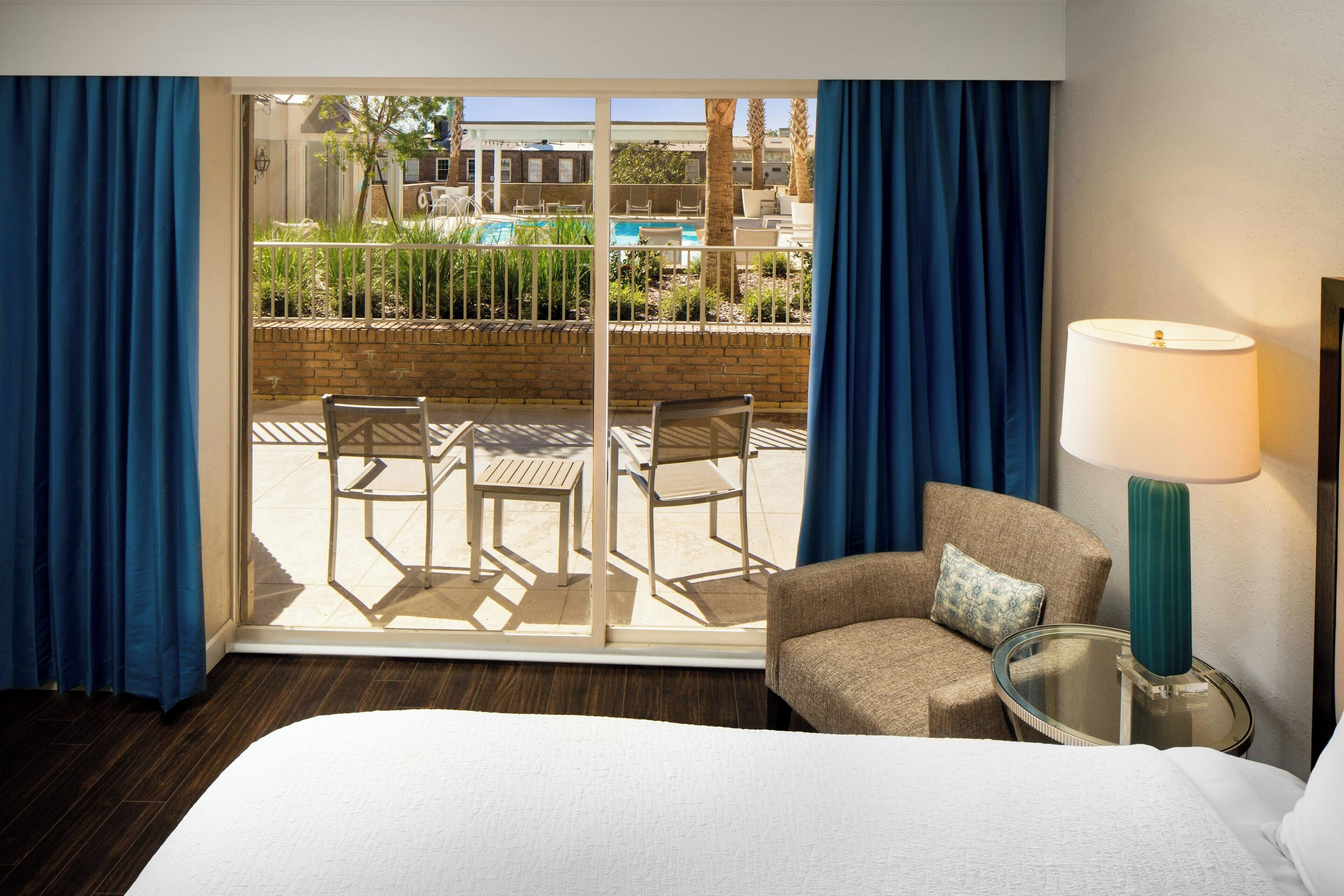 A hotel room with a bed, a chair, a table, a lamp, and a view of an outdoor pool area.