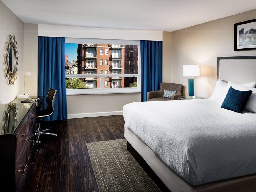 Une chambre d’hôtel avec un grand lit, un bureau avec une chaise, une télévision, une fenêtre avec des rideaux et une vue sur les bâtiments.