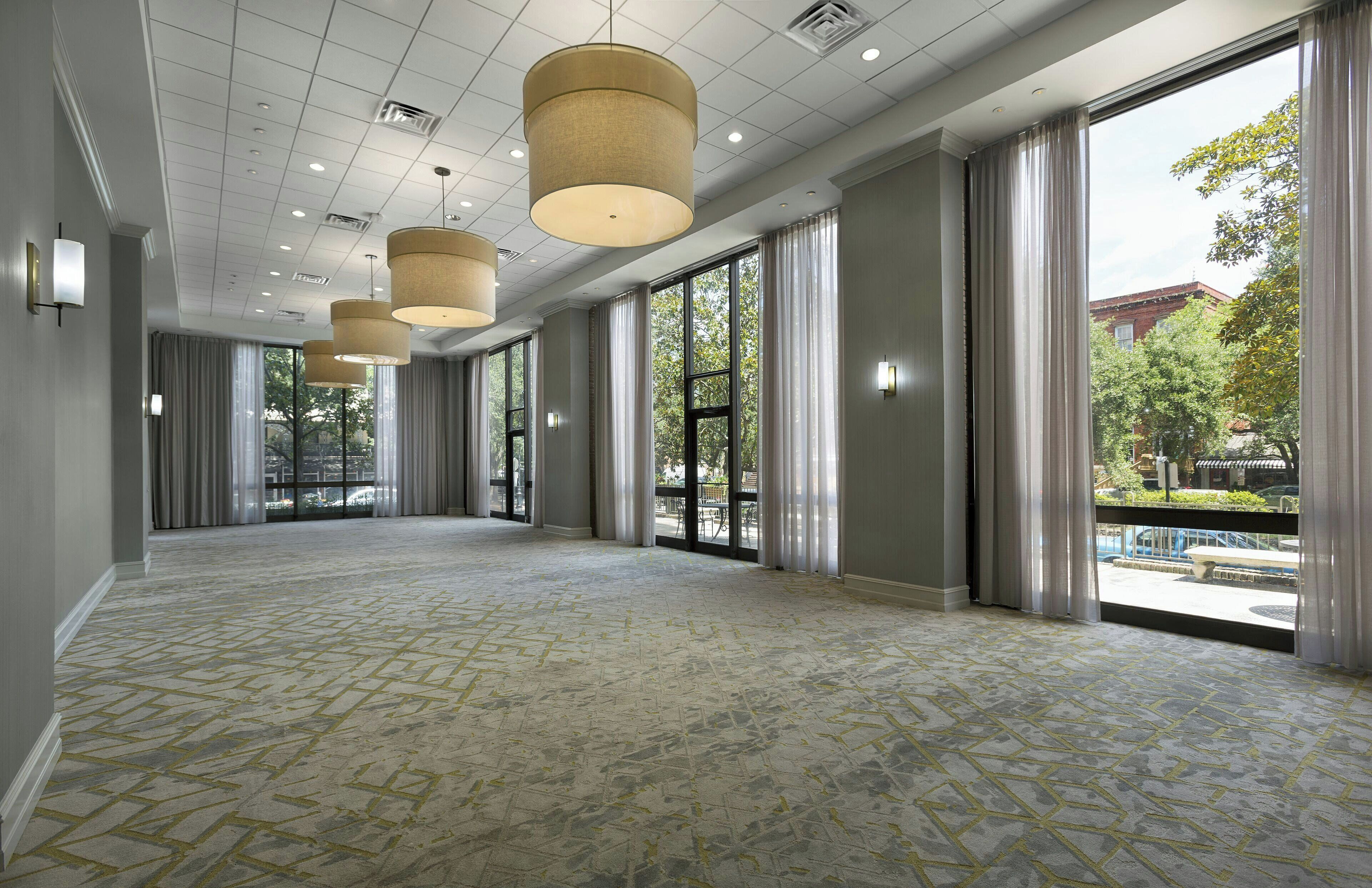A spacious hotel corridor with large windows, a patterned carpet, and hanging pendant lights.