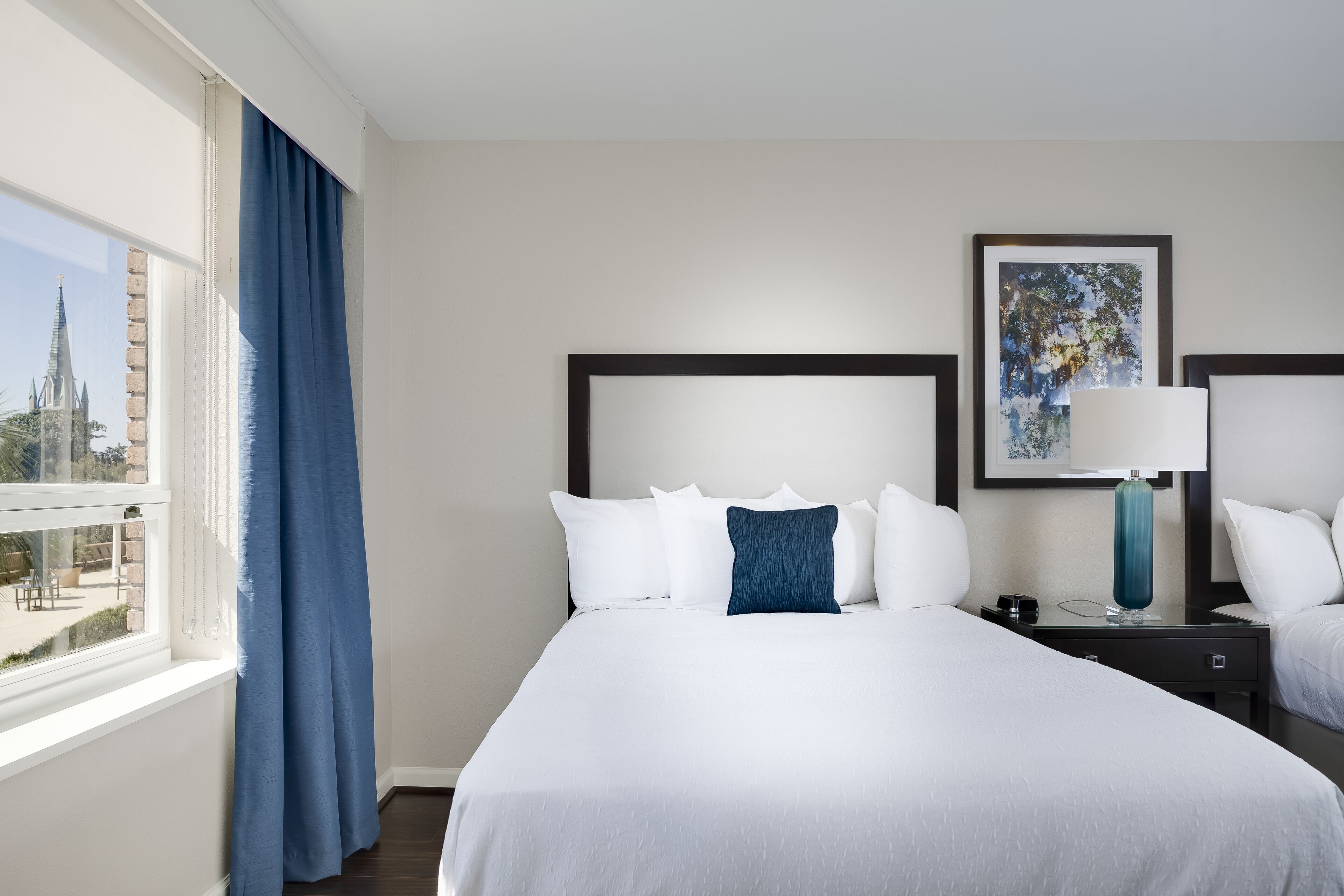 A hotel room with a large bed, a nightstand with a lamp, a framed picture on the wall, and a view of a building outside.