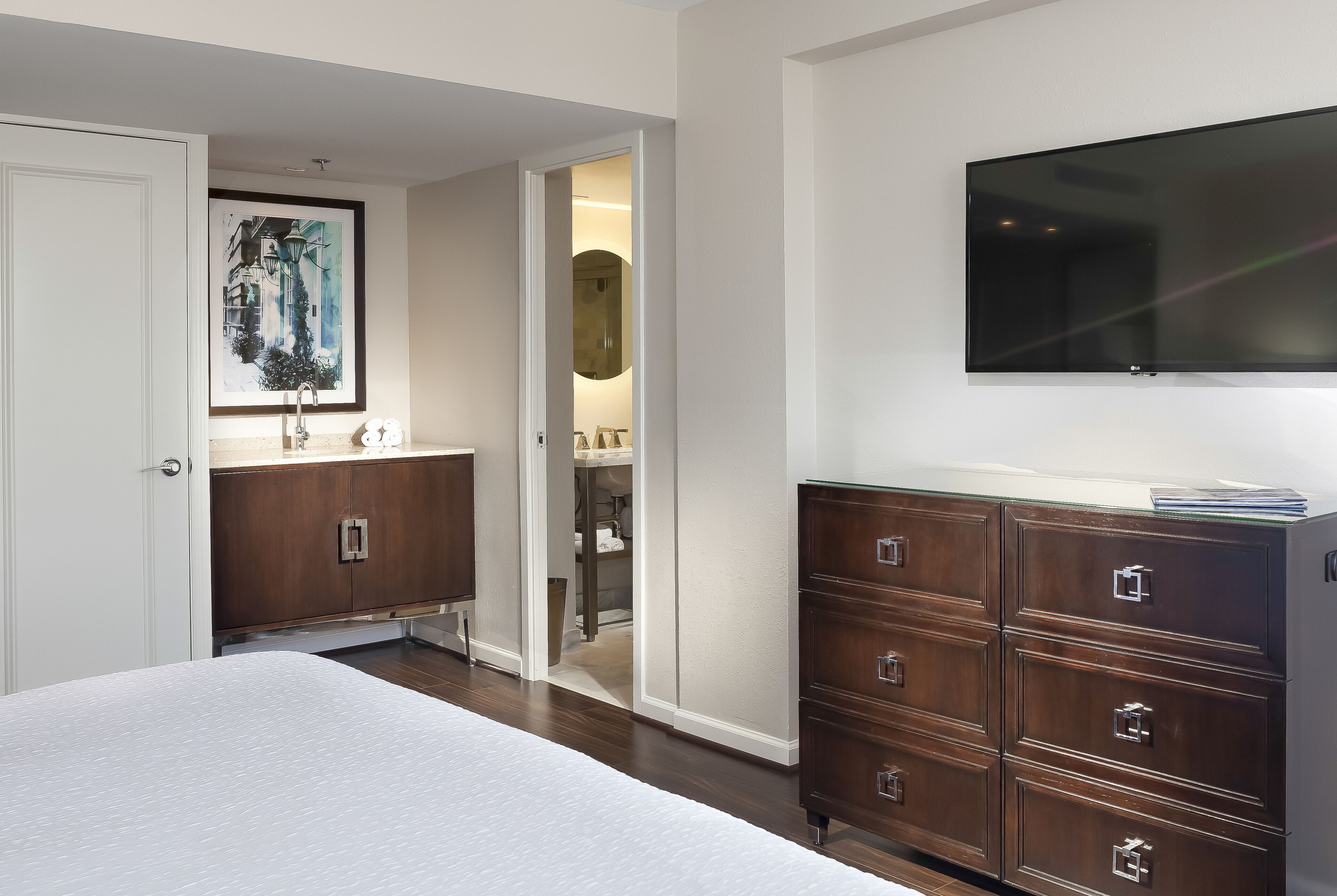 A bedroom with a bed, a dresser, a TV mounted on the wall, and a door leading to a bathroom.