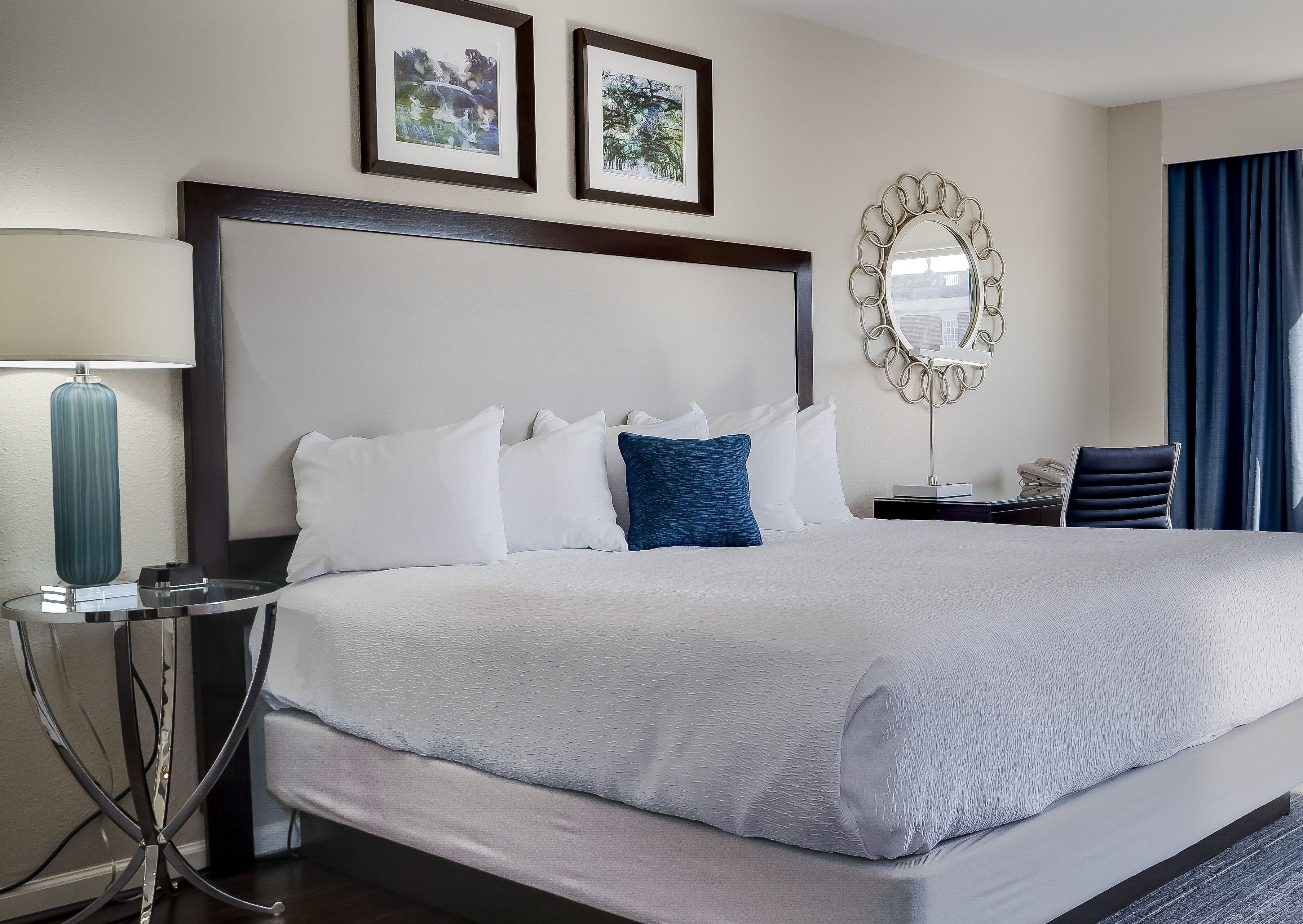A hotel room with a large bed, a bedside table with a lamp, a chair, and two framed pictures on the wall.