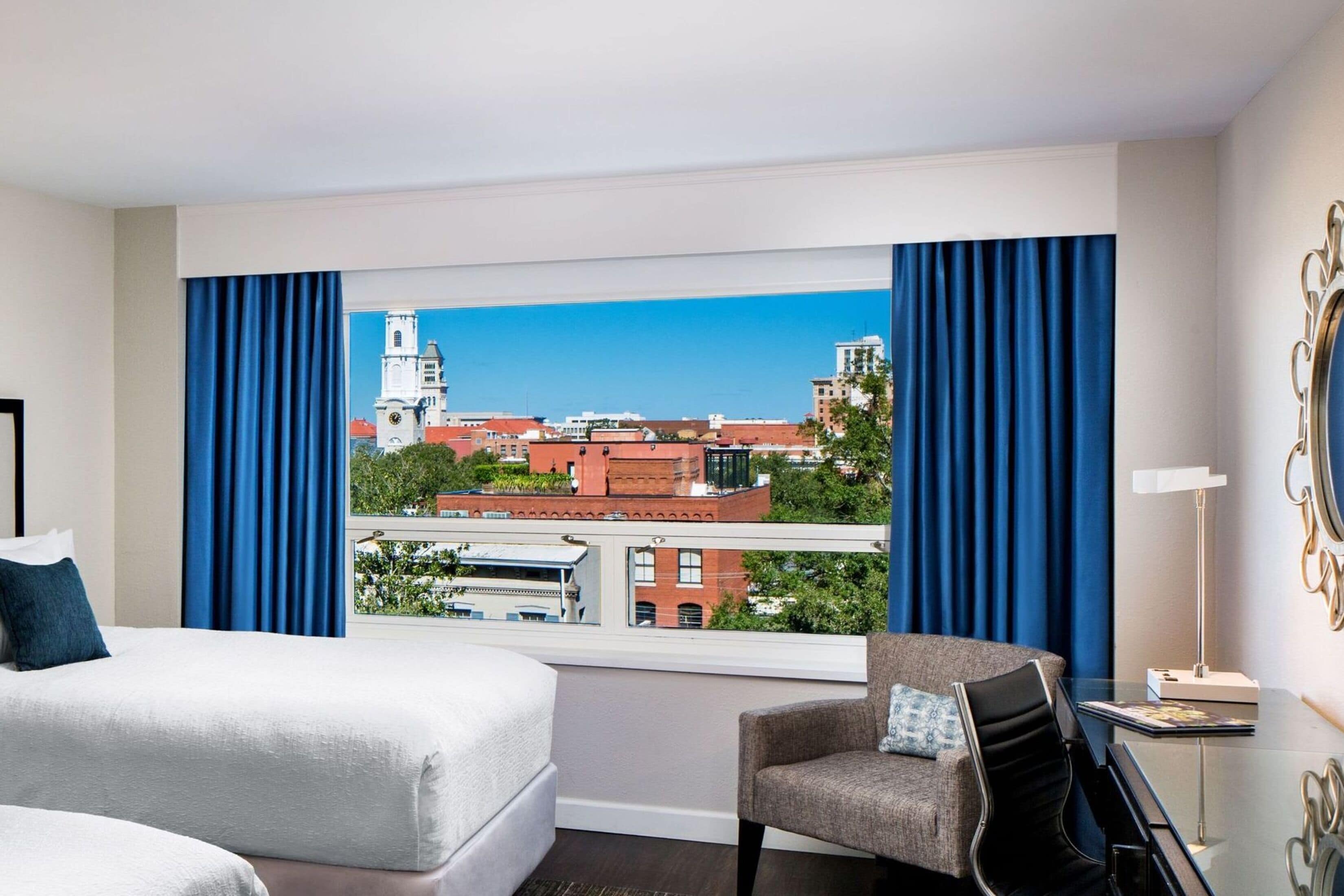 Habitación de hotel con cama, una silla, un escritorio y vistas de un paisaje urbano a través de amplios ventanales.