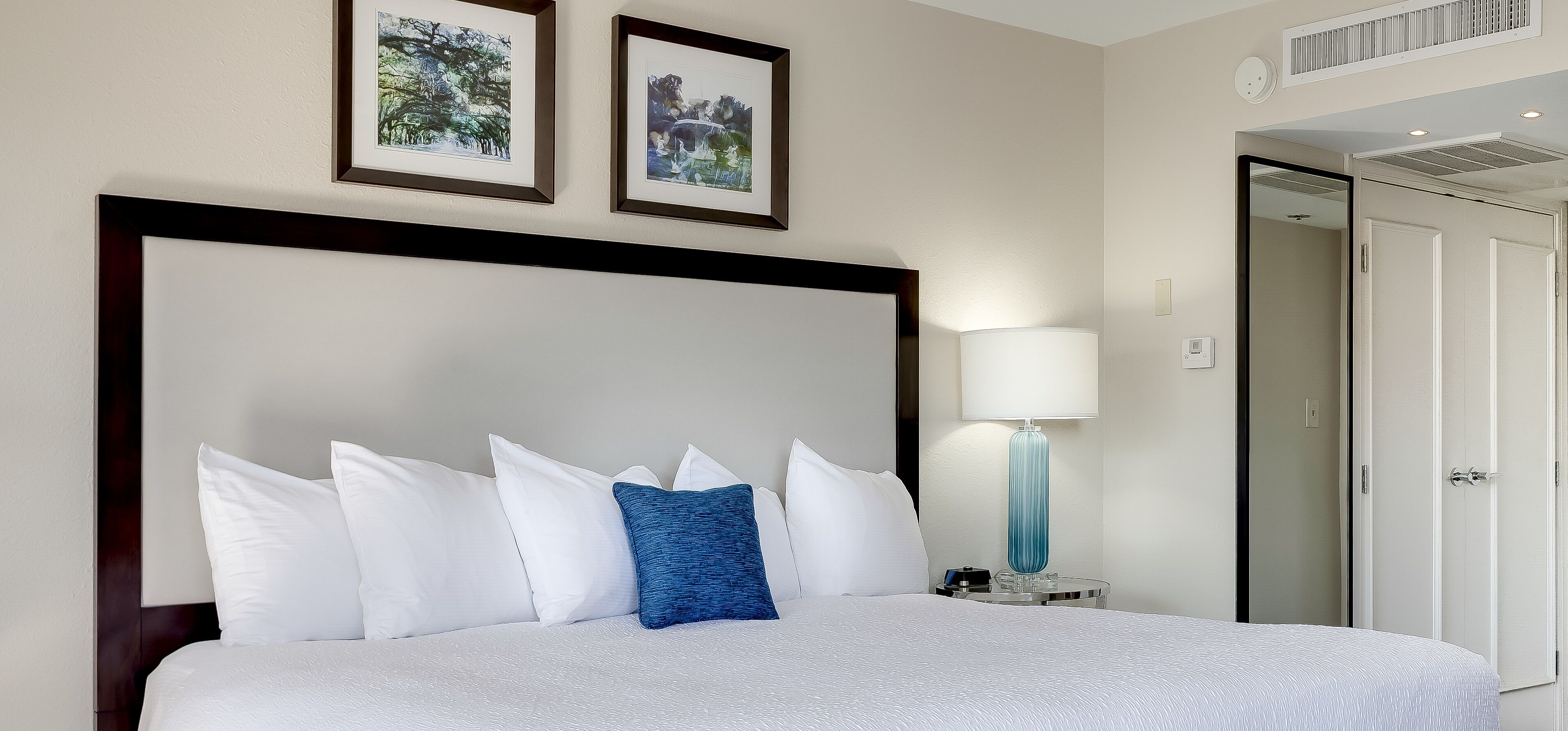 A hotel room with a large bed, white bedding, a blue accent pillow, a bedside lamp, and two framed pictures on the wall.