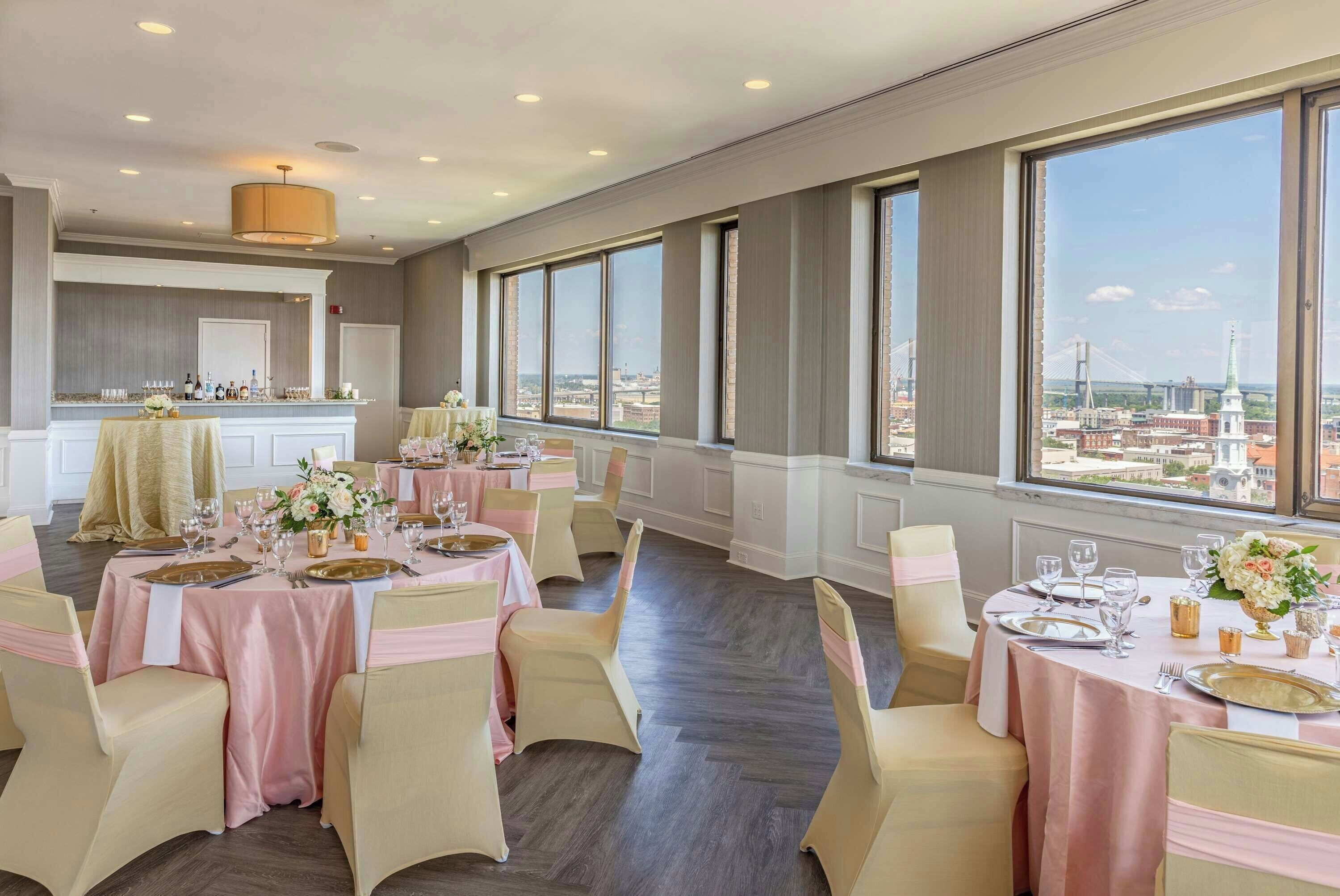 Un comedor amplio con varias mesas dispuestas para un evento formal, grandes ventanales con vistas a la ciudad y una zona de bar con mostrador.