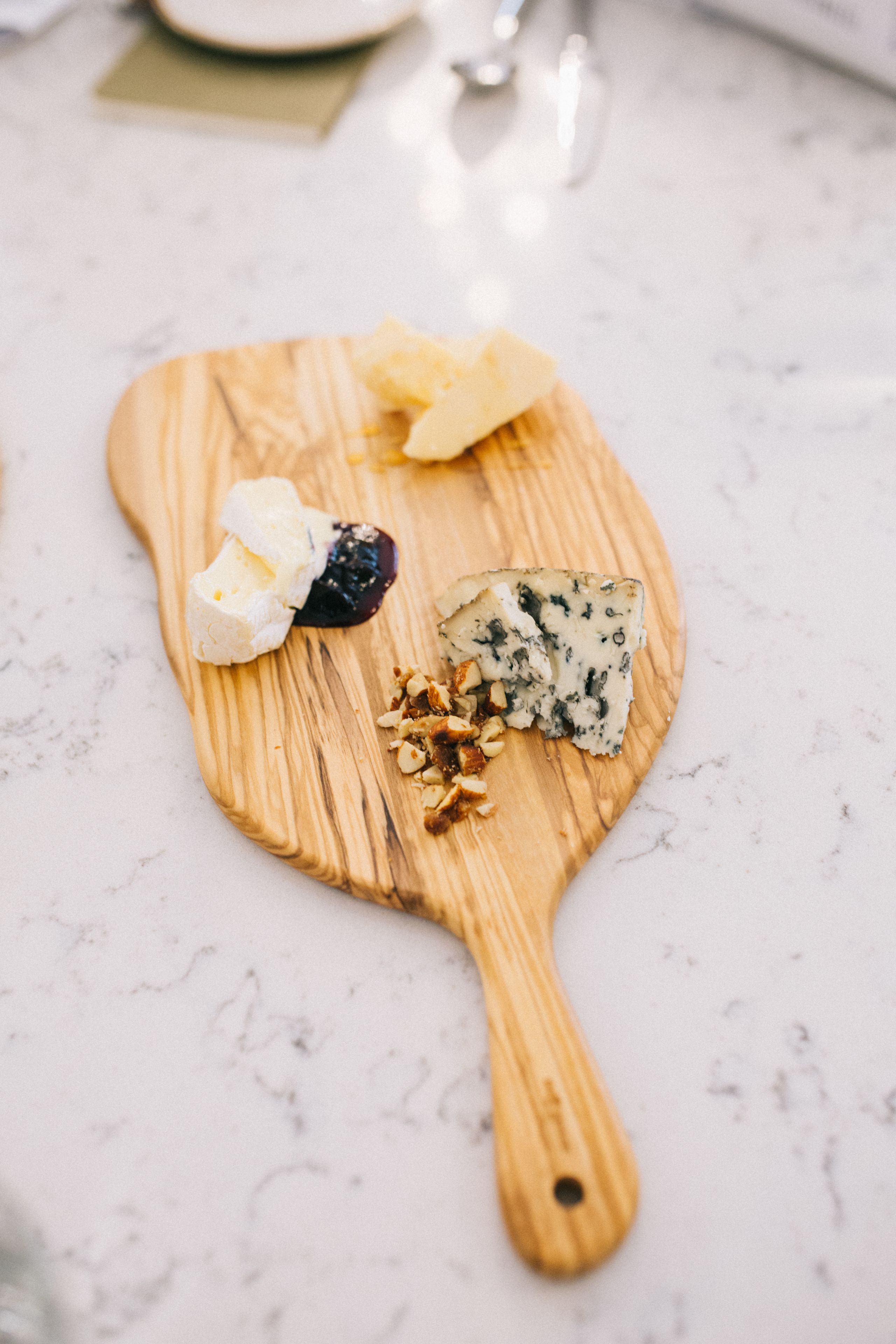 A wooden cheese board with various cheeses and nuts on a marble countertop.