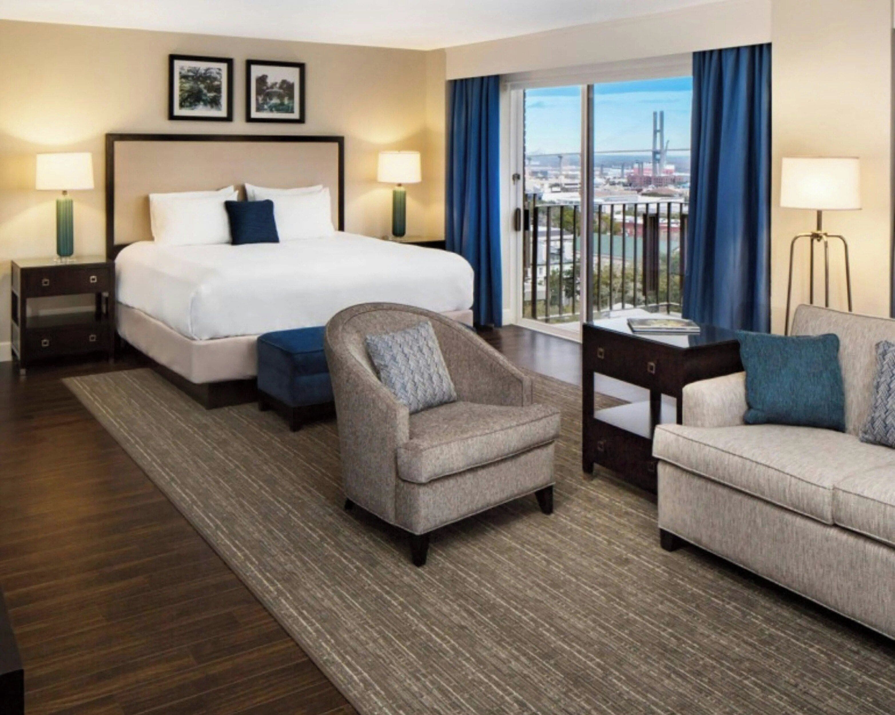 Habitación de hotel con cama, dos sillas, una mesita y vista a un paisaje urbano a través de un ventanal grande.