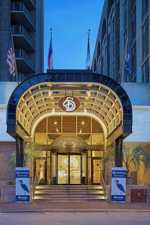 A hotel entrance with a grand archway, a large illuminated logo, and two flags. - The DeSoto (Savannah)