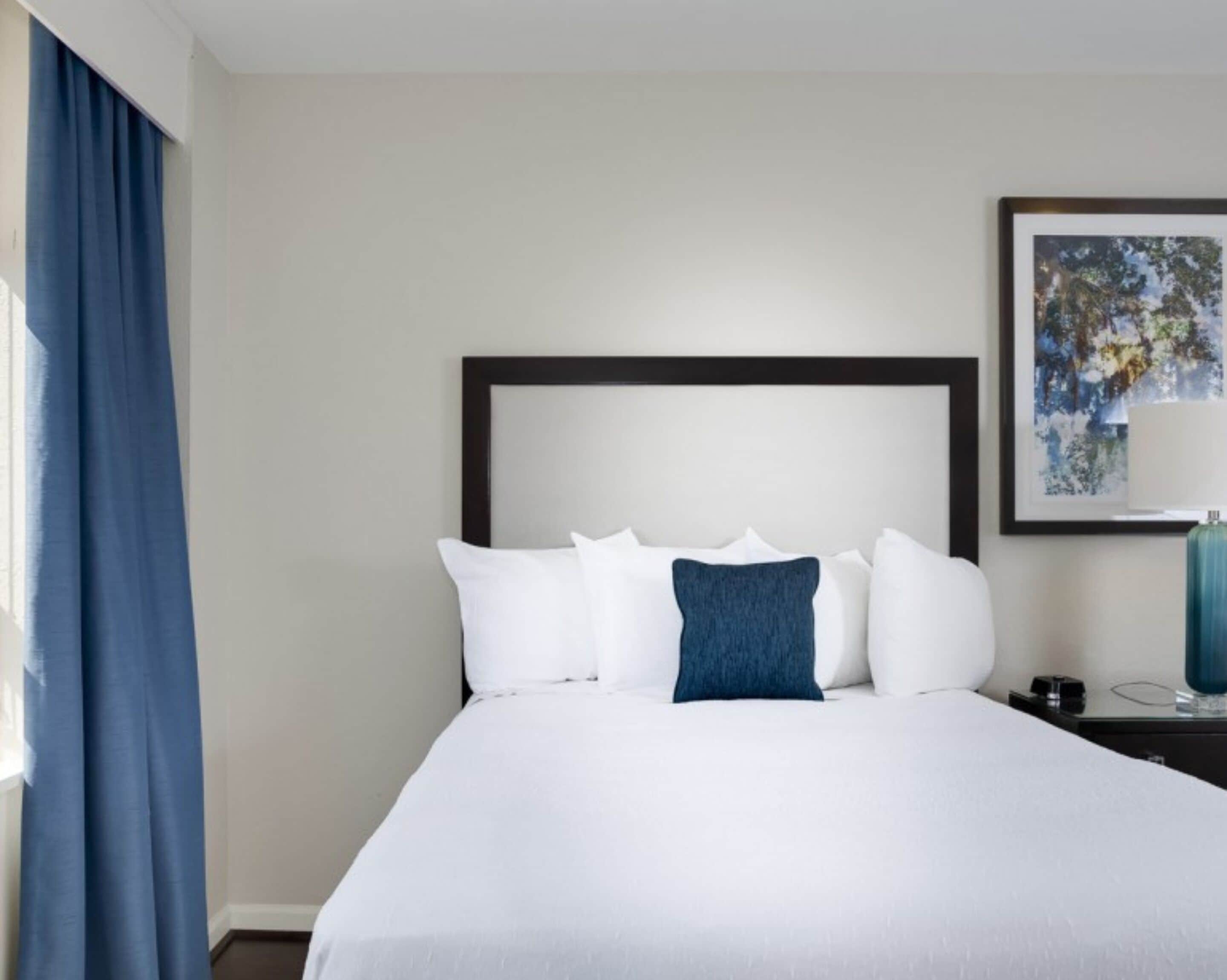 Una habitación de hotel con una cama grande, cabecera, un cuadro enmarcado en la pared y una mesita de noche con lámpara.