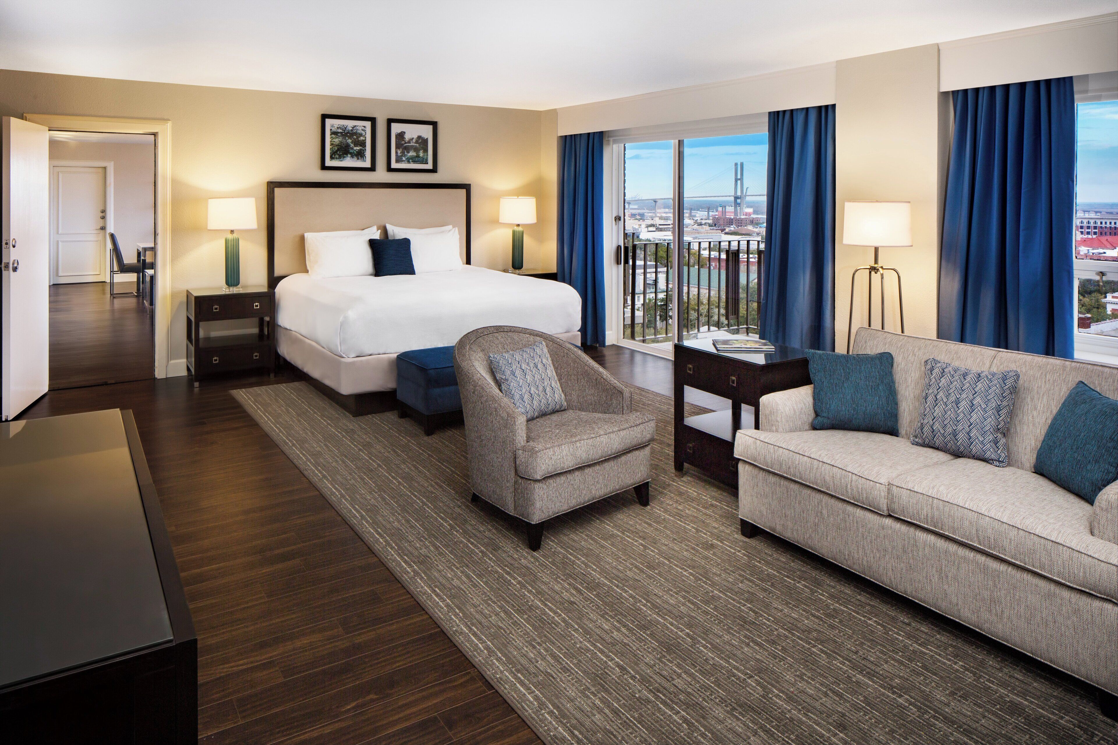 Une chambre d’hôtel avec un grand lit, deux fauteuils, un canapé, un bureau et une vue sur le paysage urbain.
