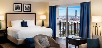 A hotel room with a large bed, two bedside lamps, a chair, a small table, and a view of a cityscape with a bridge.