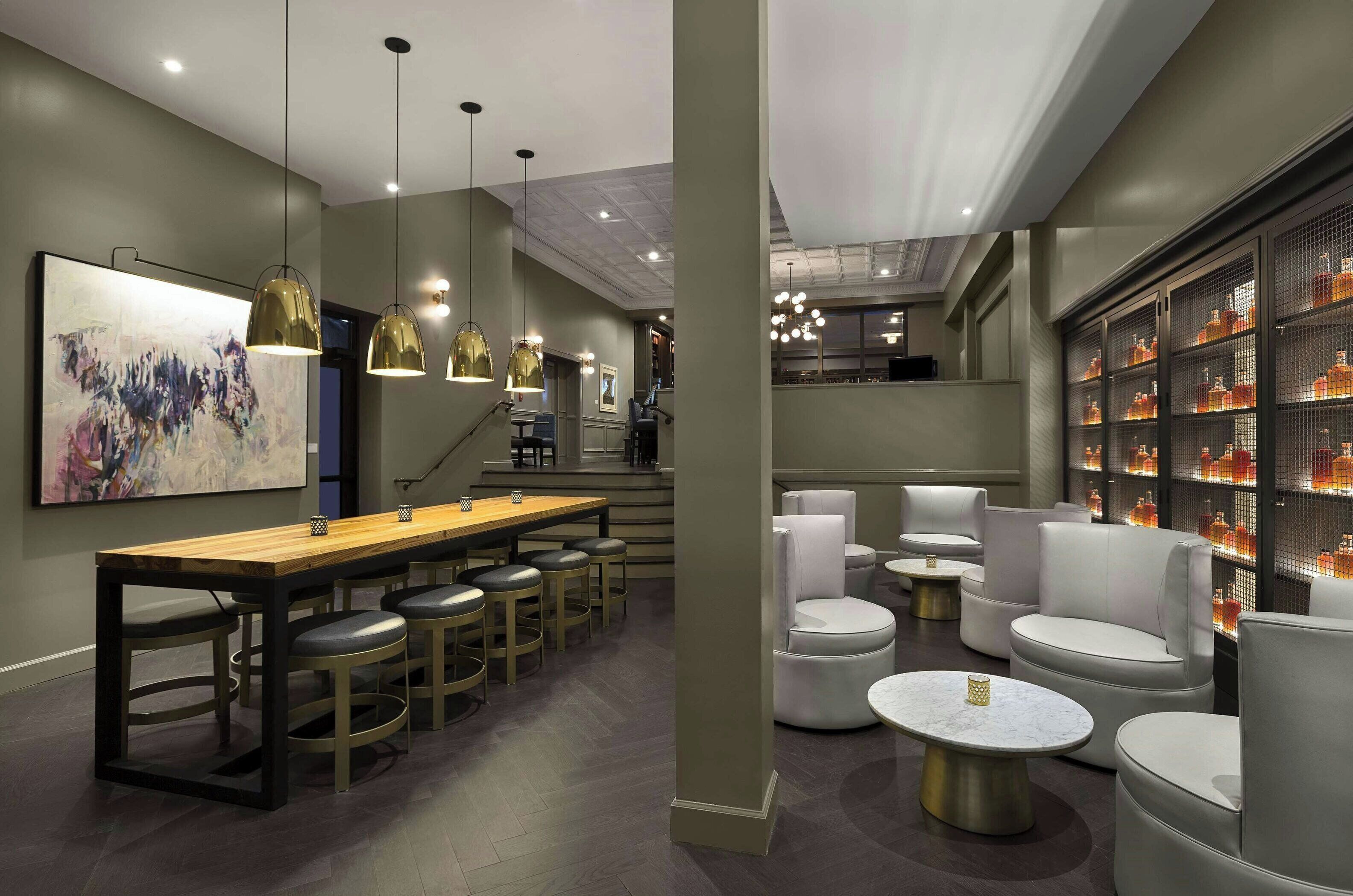 Un espace bar moderne doté d’un long comptoir en bois, de nombreux tabourets, d’un éclairage suspendu et d’un coin salon avec des tables rondes et des chaises rembourrées.