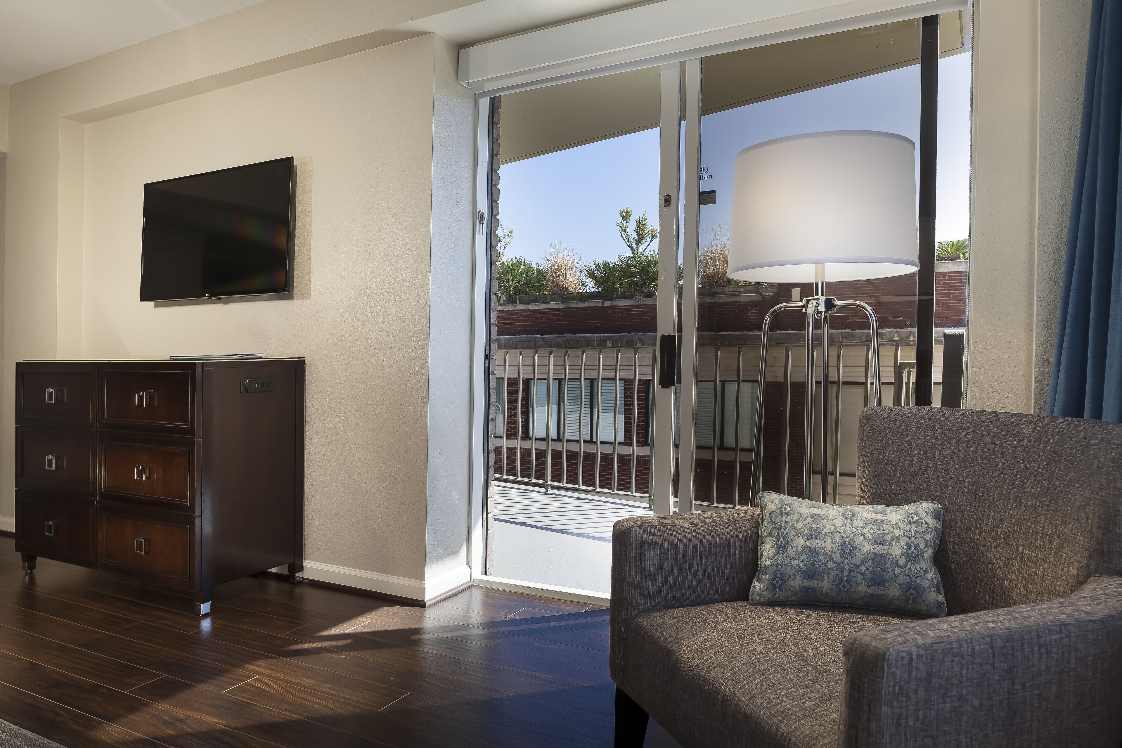 A modern living room with a dark wood dresser, a flat-screen TV mounted on the wall, a gray sofa with a patterned pillow, and a large sliding glass door leading to an outdoor balcony with a white lamp.