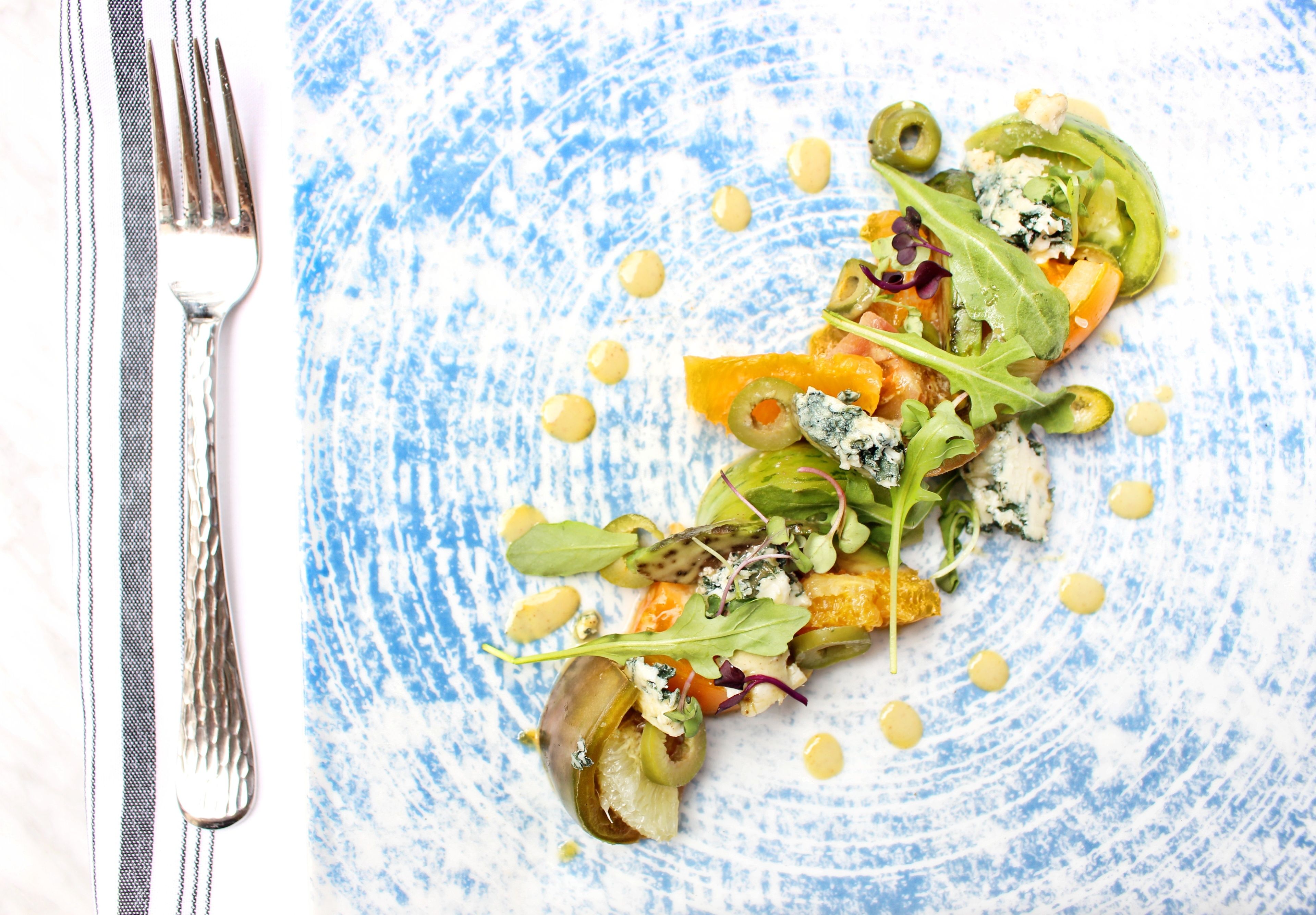 A plate of food with various vegetables and a blue sauce, accompanied by a fork and knife on a blue textured plate.