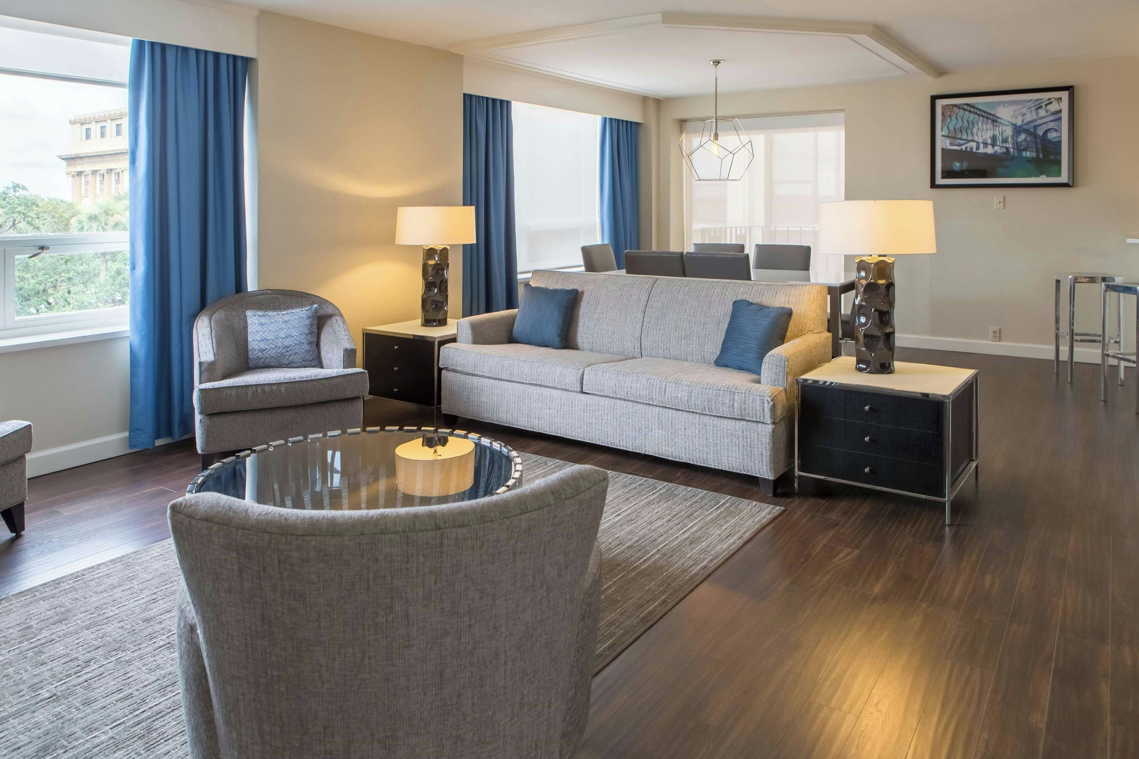 A modern living room with a grey sofa, armchairs, a glass coffee table, and a dining area with chairs.