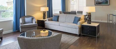 A modern living room with a grey sofa, armchairs, a glass coffee table, and a dining area with chairs.