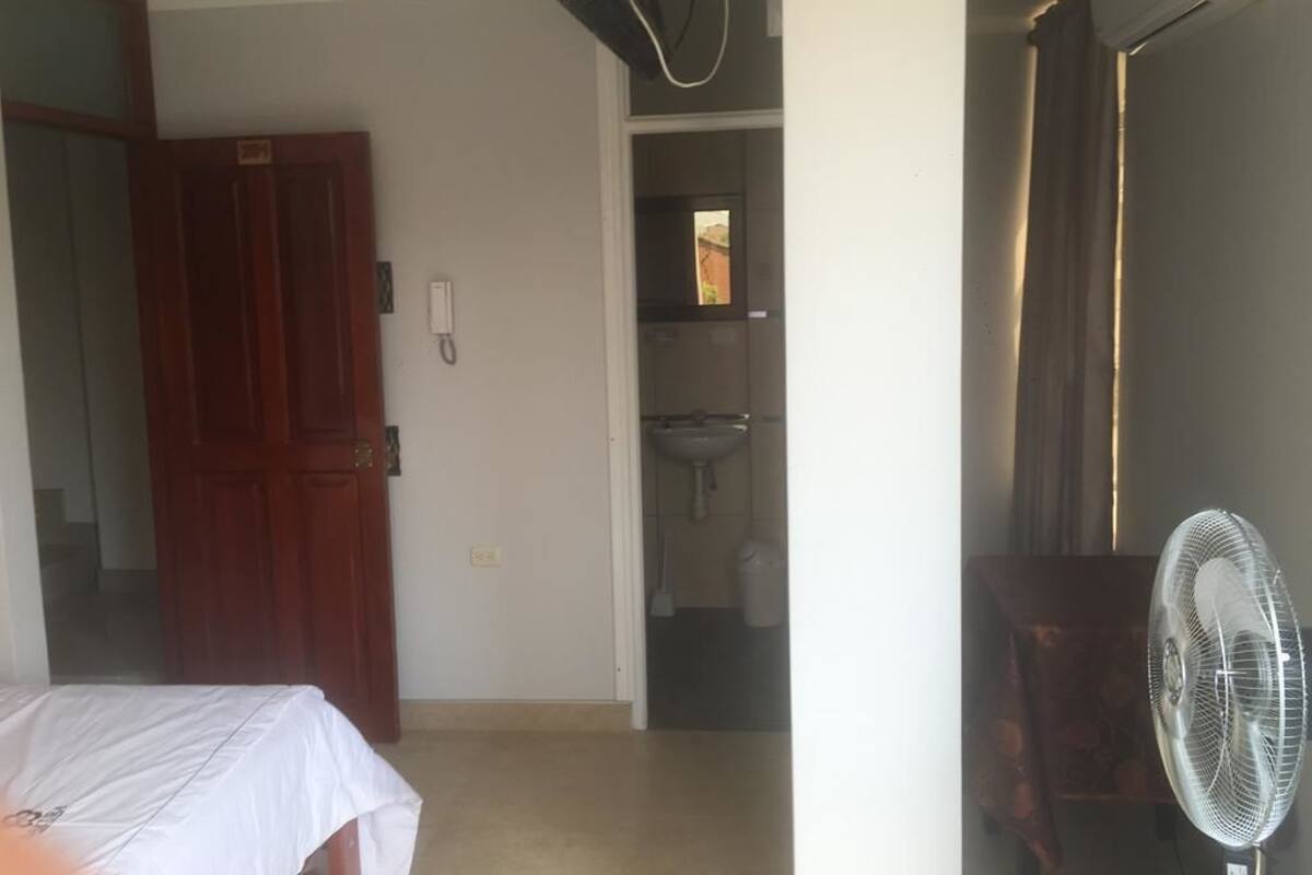 Habitación de hotel con cama, puerta de madera, baño con lavamanos y un ventilador de pie.