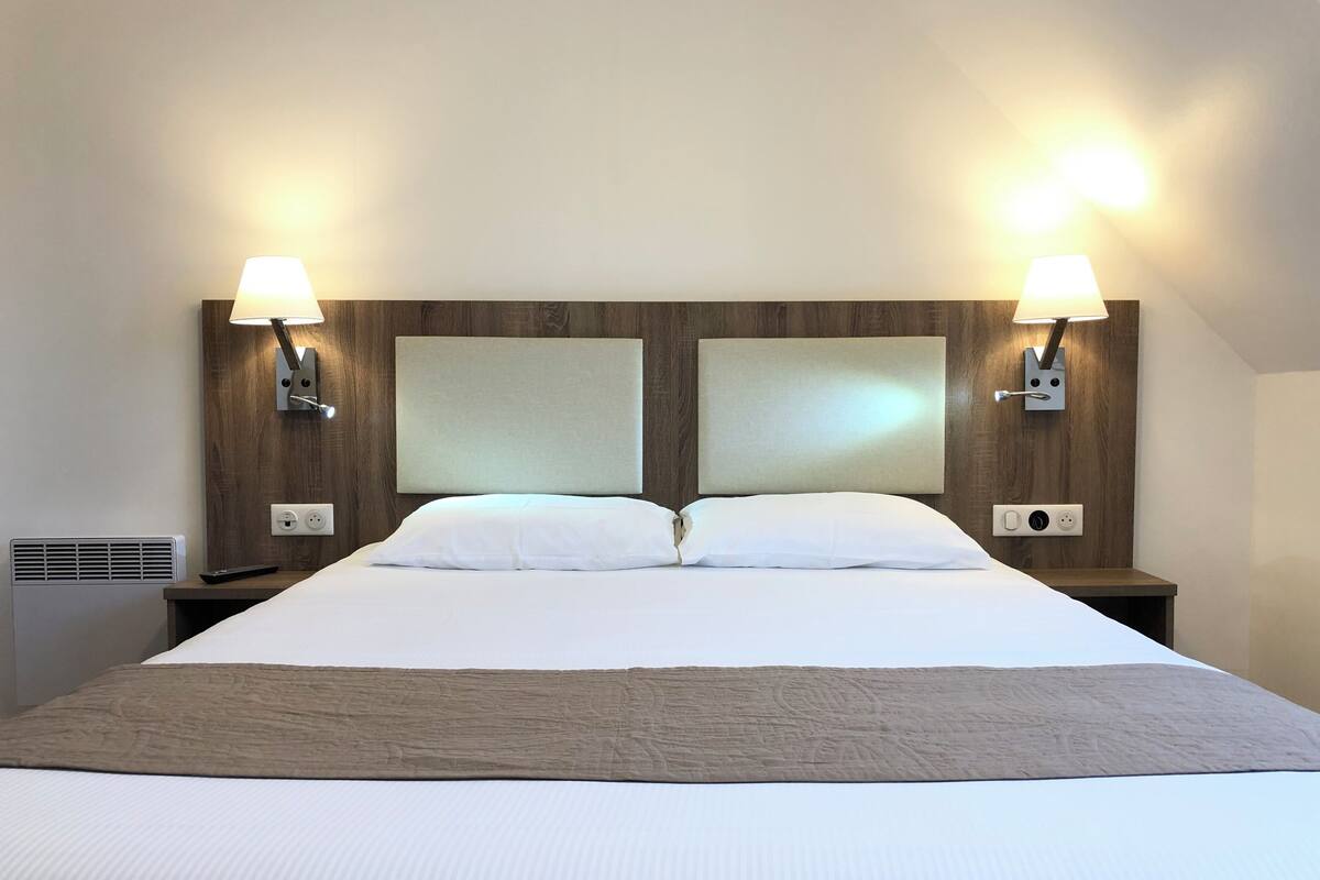 Una habitación de hotel con una cama grande, dos lámparas de noche, un cabecero y un enchufe.