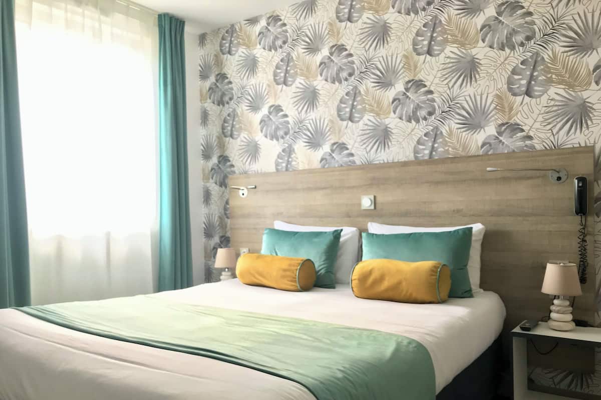 Habitación de hotel con una cama, dos almohadas, una mesita de noche con una lámpara y una pared con diseño de hojas.