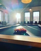 A pool table with a label reading "Billiards BENARD" in a room with large windows and a hanging light fixture.