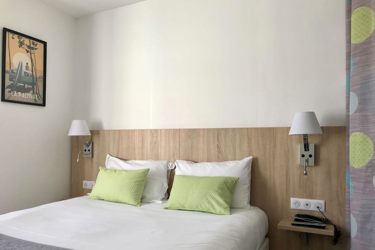 Una habitación de hotel con cabecera de madera, dos lámparas de noche, una cama con almohadas blancas y verdes, una mesita de noche pequeña con una lámpara y un enchufe.