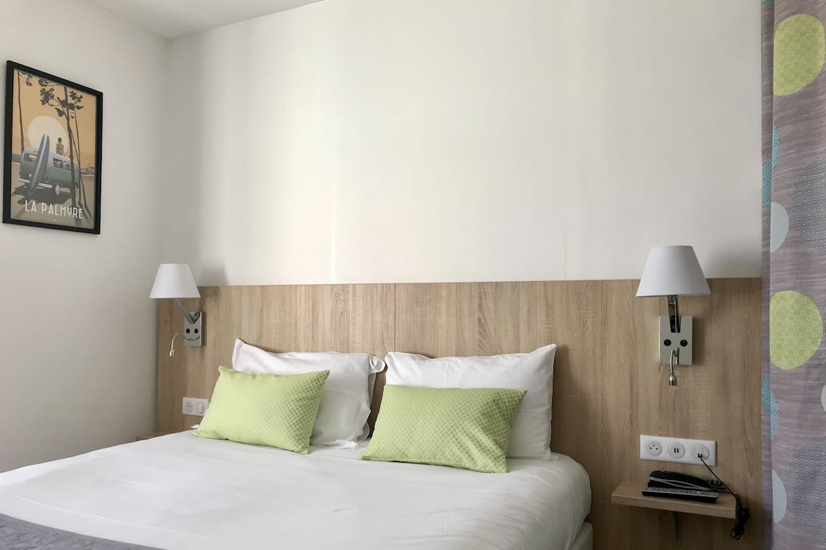 Una habitación de hotel con cabecera de madera, dos lámparas de noche, una cama con almohadas blancas y verdes, una mesita de noche pequeña con una lámpara y un enchufe.