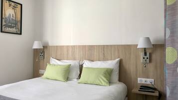 Une chambre d’hôtel avec une tête de lit en bois, deux lampes de chevet, un lit avec des oreillers blancs et verts, une petite table de chevet avec une lampe et une prise de courant.