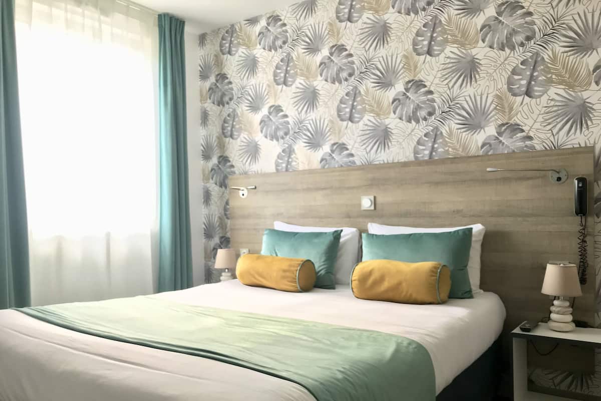 Habitación de hotel con una cama, dos almohadas, una mesita de noche con una lámpara y una pared con diseño de hojas.