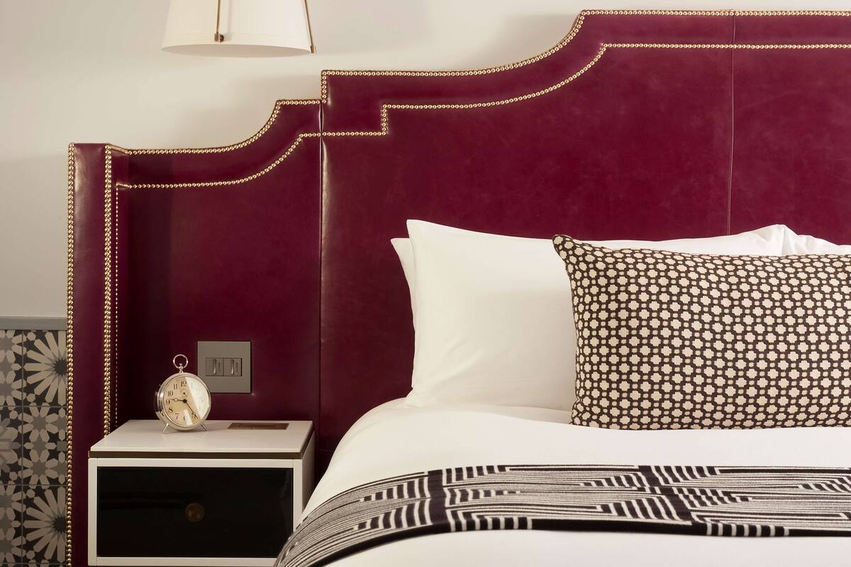 A bed with a patterned pillow, a bedside table with a clock, and a wall with a geometric tile pattern.