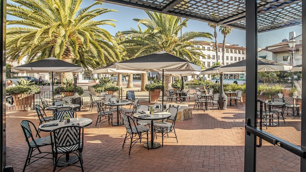 An outdoor patio with tables and chairs, palm trees, and a view of buildings in the background. - Hotel Californian (Santa Barbara)