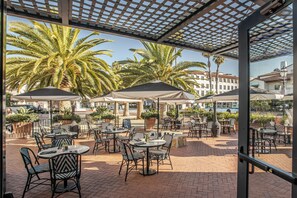 An outdoor patio with tables and chairs, palm trees, and a view of buildings in the background. - Hotel Californian (Santa Barbara)