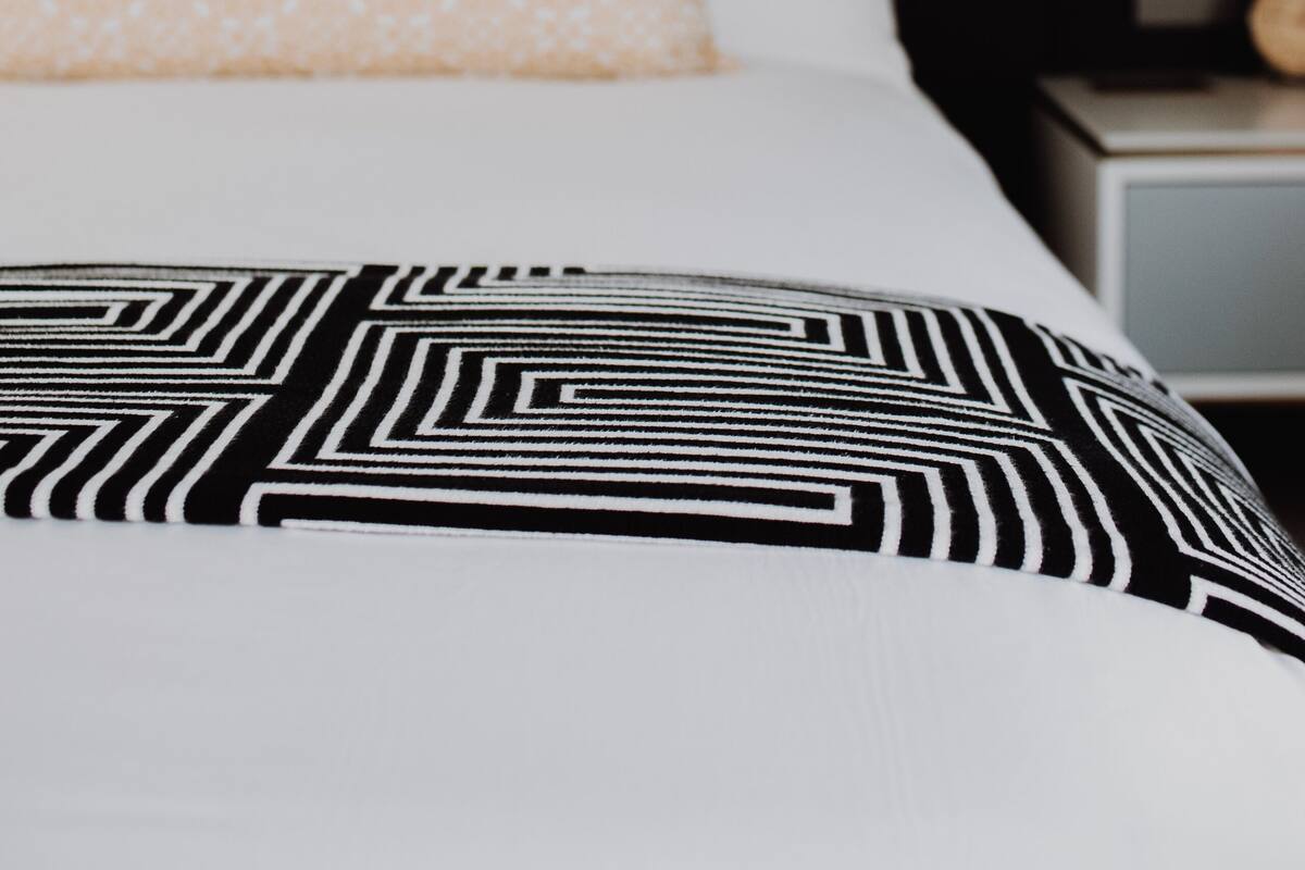 A bed with a black and white patterned bedspread, a white pillow with a light pattern, and a dark headboard.