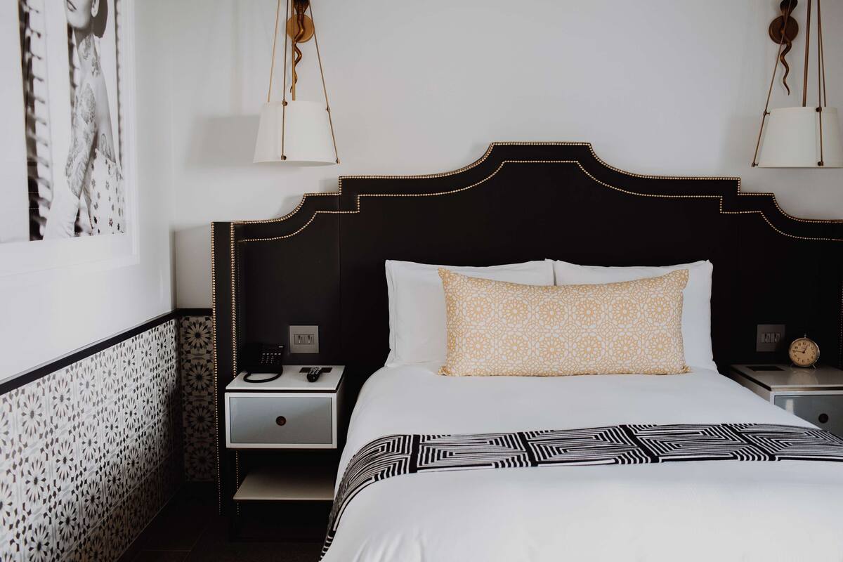 A hotel room with a black headboard, white bedding, patterned pillows, a bedside table with a lamp, and a clock.