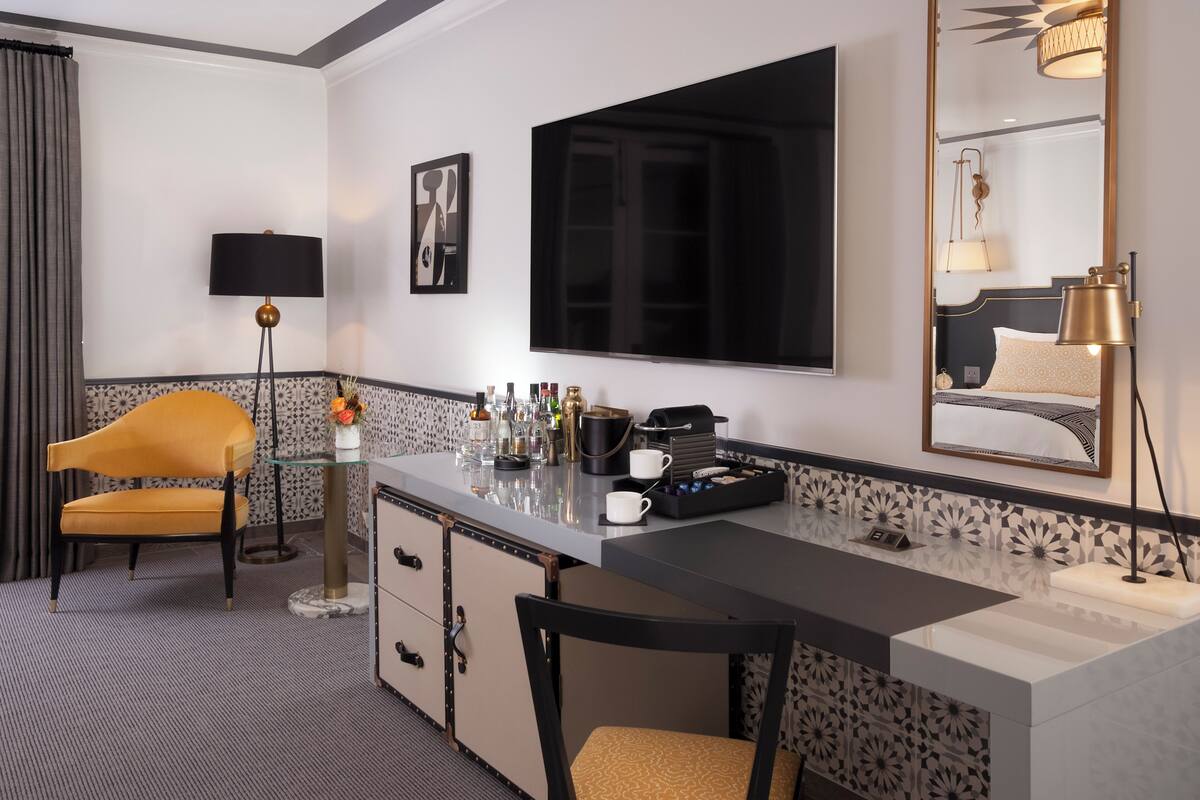 A hotel room with a minibar, a desk, a chair, a television, a large mirror, and a patterned wall.