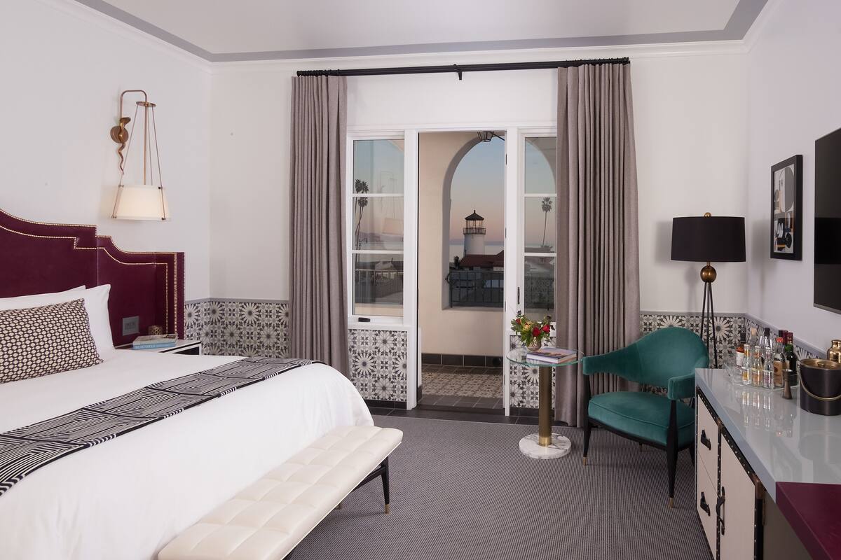 A hotel room with a large bed, a desk with a chair, a small table with a lamp, and a view of a building through the window.