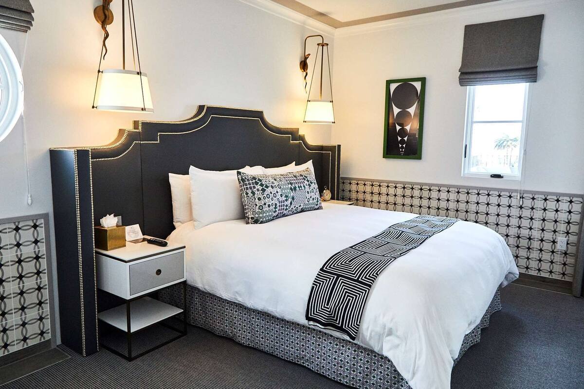 A hotel room with a large bed, a nightstand, a wall-mounted lamp, a framed picture, and a window with a view.